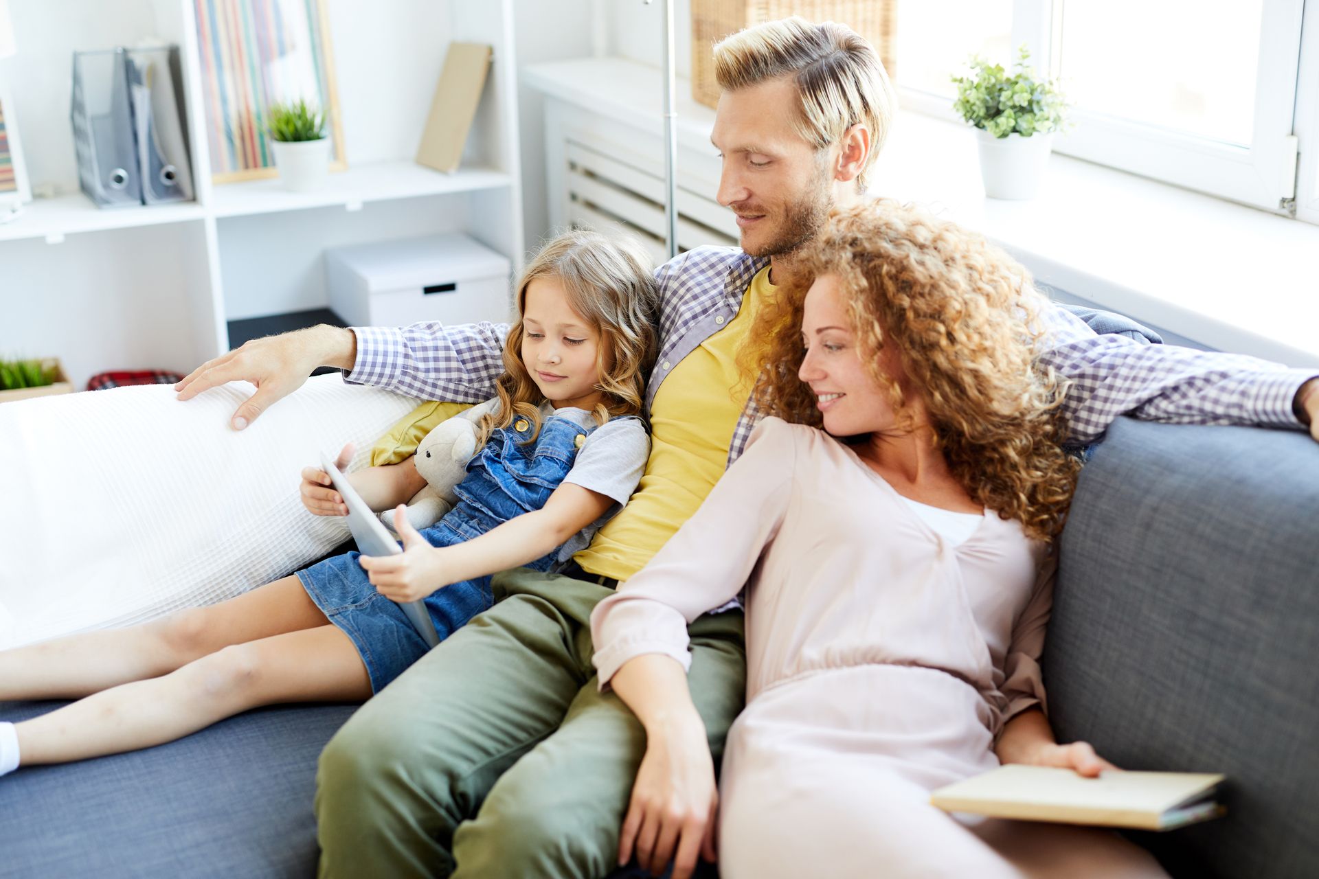 Family of three on a sofa, daughter looking at a tablet, parents smiling and looking at her.