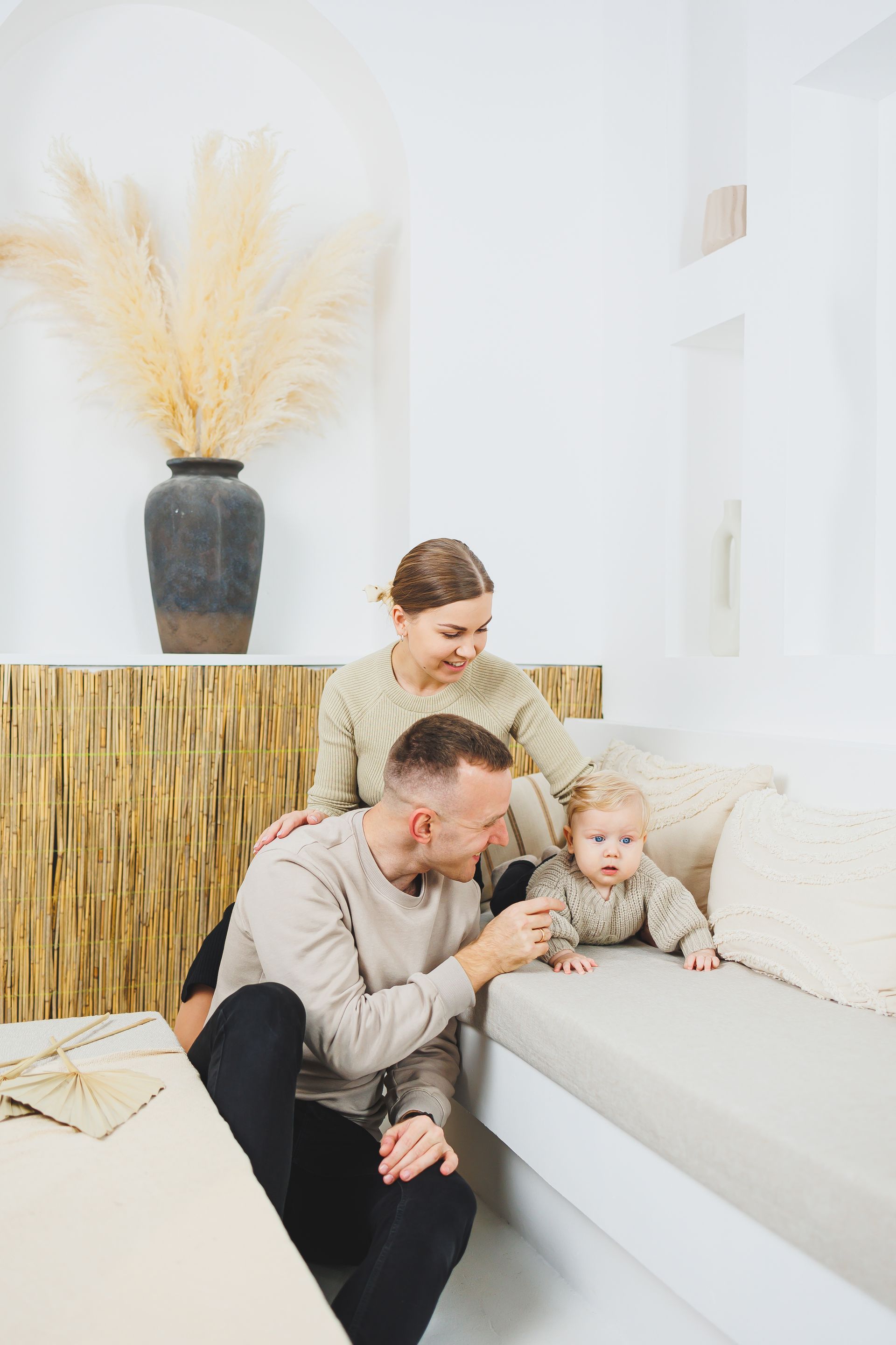 Family in neutral-toned room. Father and mother with baby, touching the baby on a bench.