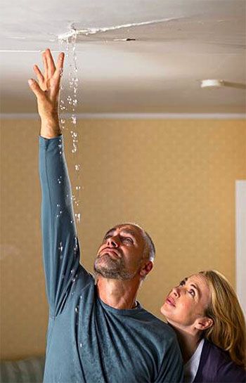Two people inspect a ceiling water leak, with water dripping down from a damaged spot.