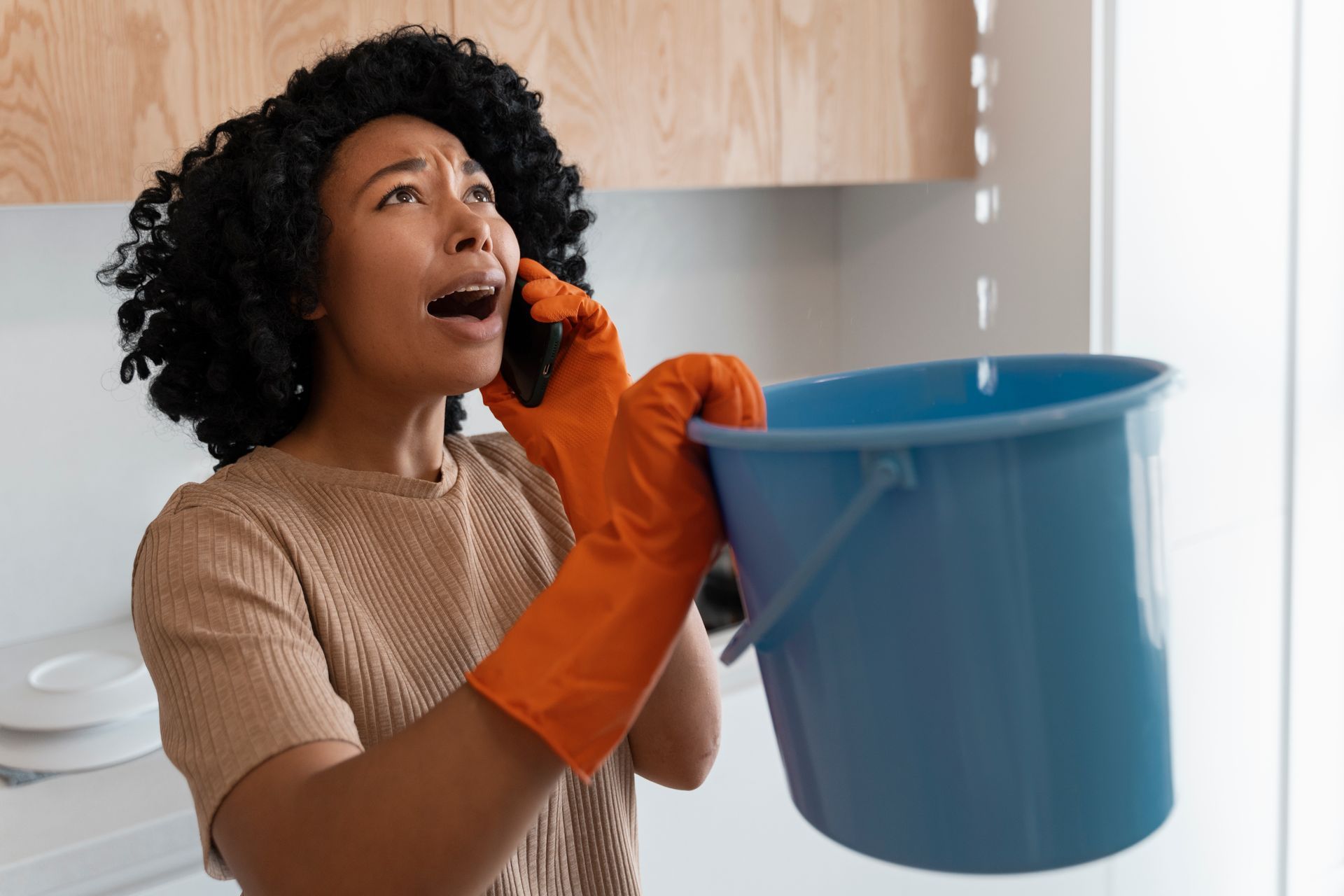 Person in orange gloves holding a blue bucket while talking on a phone in a kitchen.