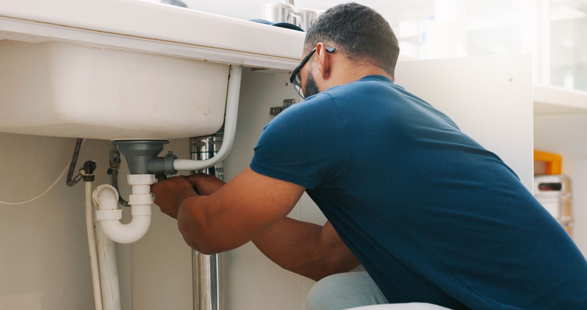Person crouching under a sink, adjusting white plumbing pipes with tools in a kitchen cabinet