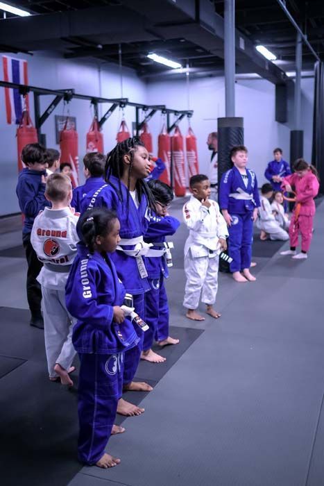 Children in martial arts uniforms standing in a gym, attentively watching a demonstration.