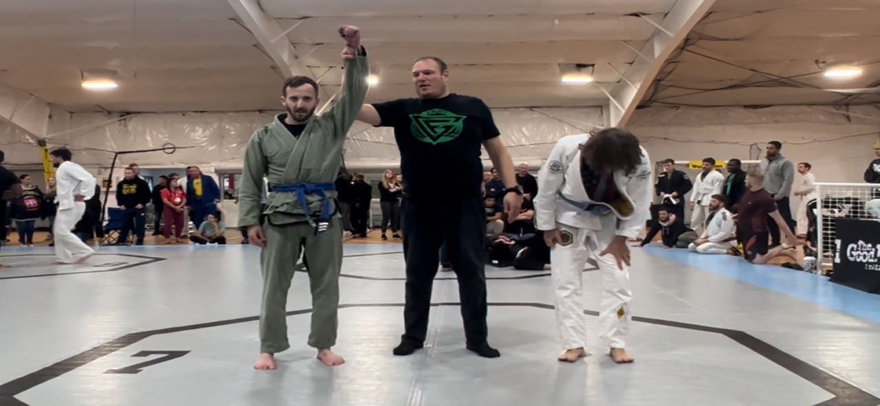 Jiu-Jitsu match: Winner in green gi raises arm as opponent in white gi bows. Referee stands between them in a gym.