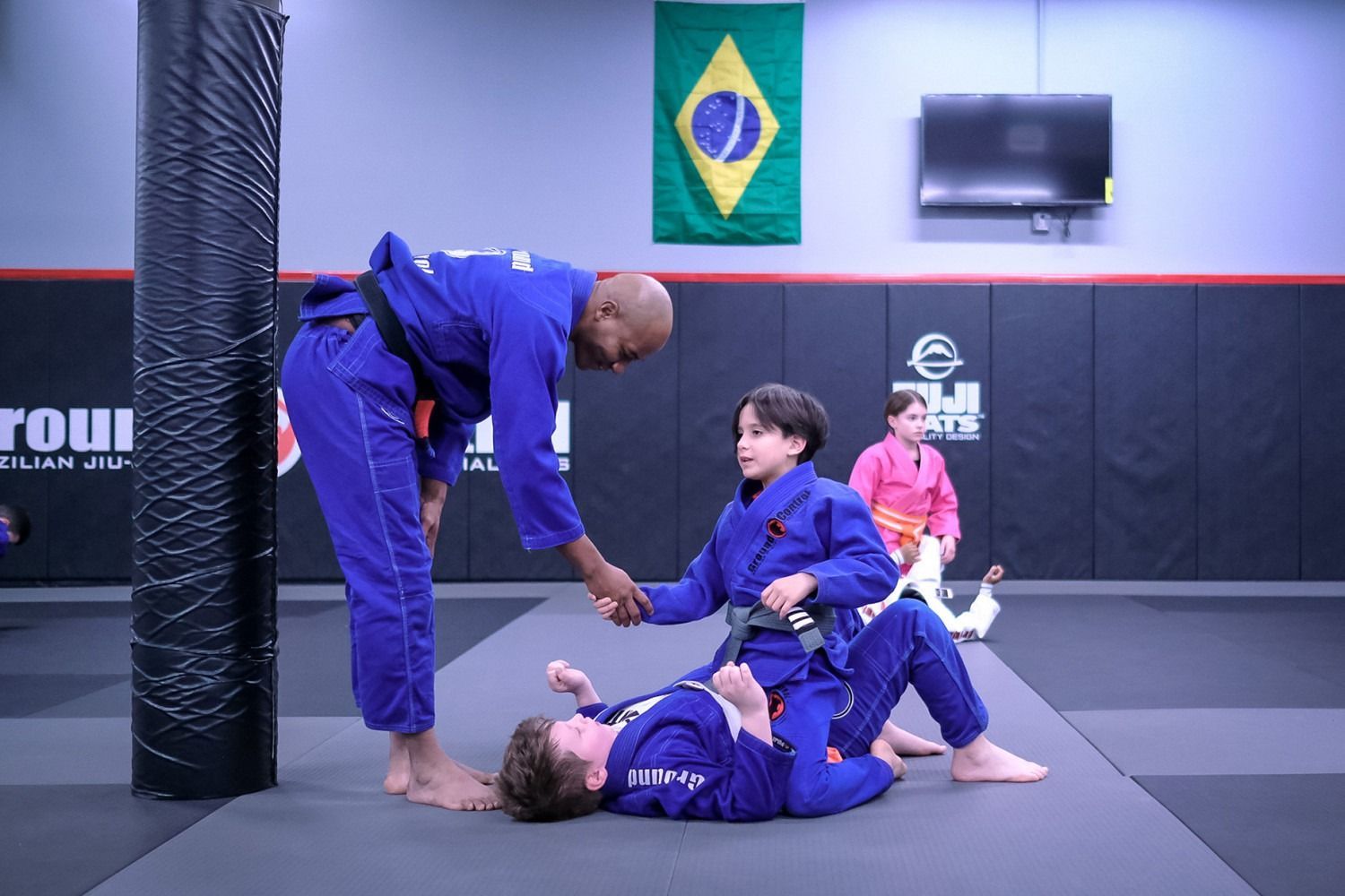 Instructor shaking hands with a student in blue gi, other child on the mat. Brazilian Jiu-Jitsu gym.