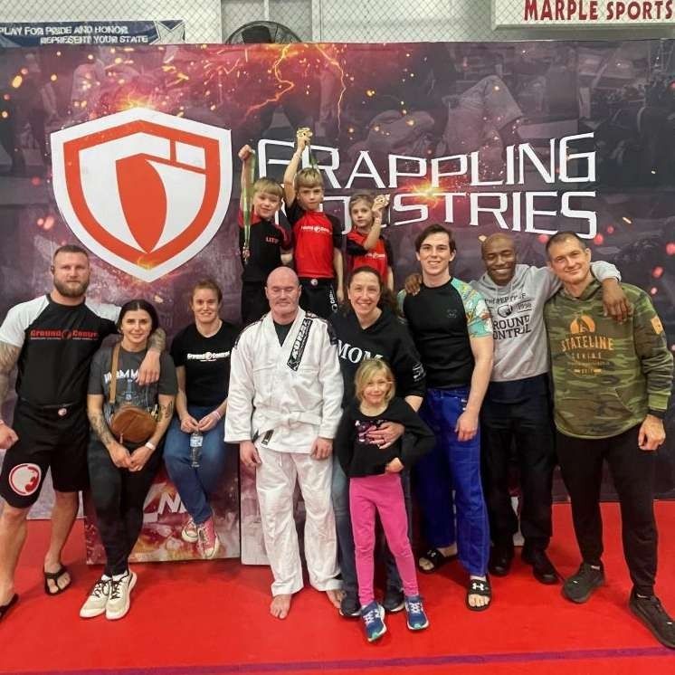 Group photo at Grappling Industries event; diverse group smiling, some children; banner backdrop.