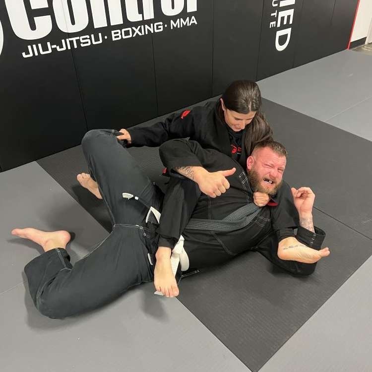 A person in a black gi, in a grappling hold by a young person in a black gi on a mat, with a gym background.