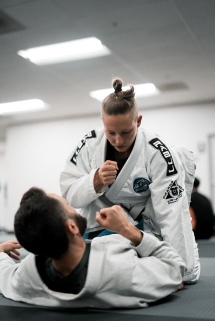 Woman in white gi kneeling over a man on the mat, practicing Jiu-Jitsu.