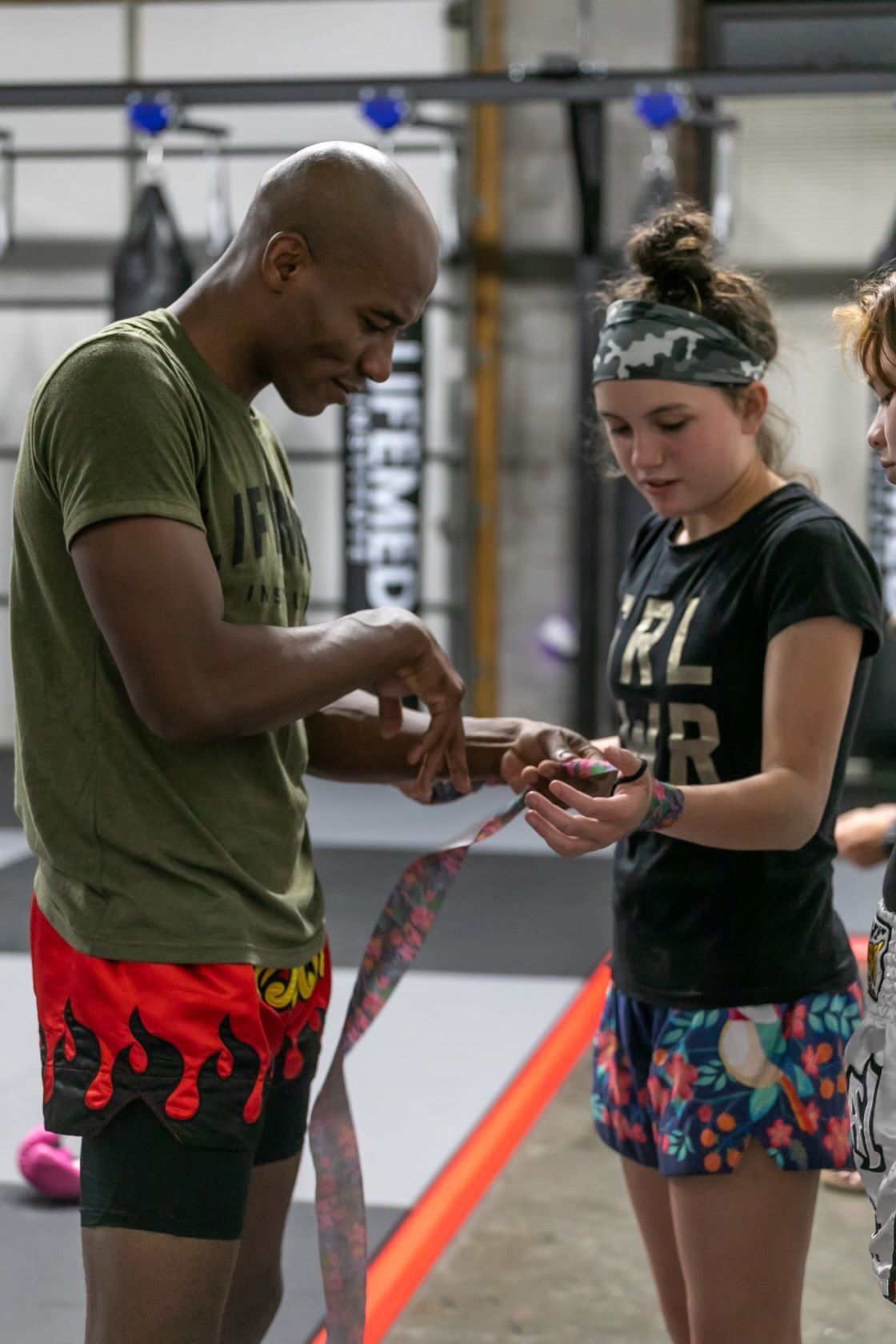 Man wrapping hand, helping woman at gym. Man wears olive shirt, red shorts; woman wears black shirt, floral shorts.