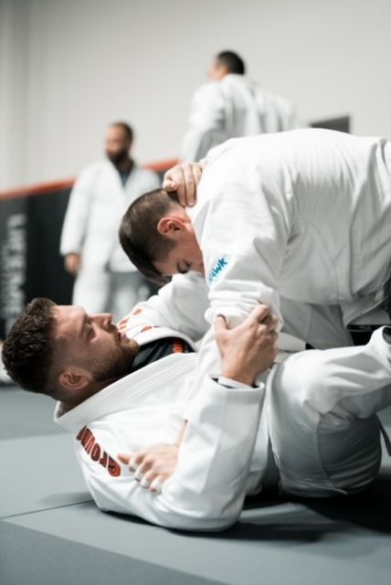 Two men in white gis practice Brazilian Jiu-Jitsu. One on the ground, the other atop. Other people in background.