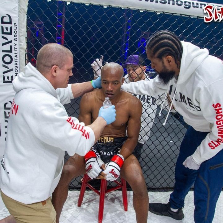 A fighter, seated, being attended to in a cage. Men in white hoodies tend to him, with a water bottle and gloves.