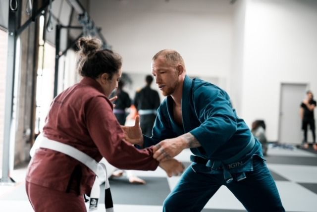 Woman in maroon gi and man in blue gi practicing Jiu-Jitsu in a gym.
