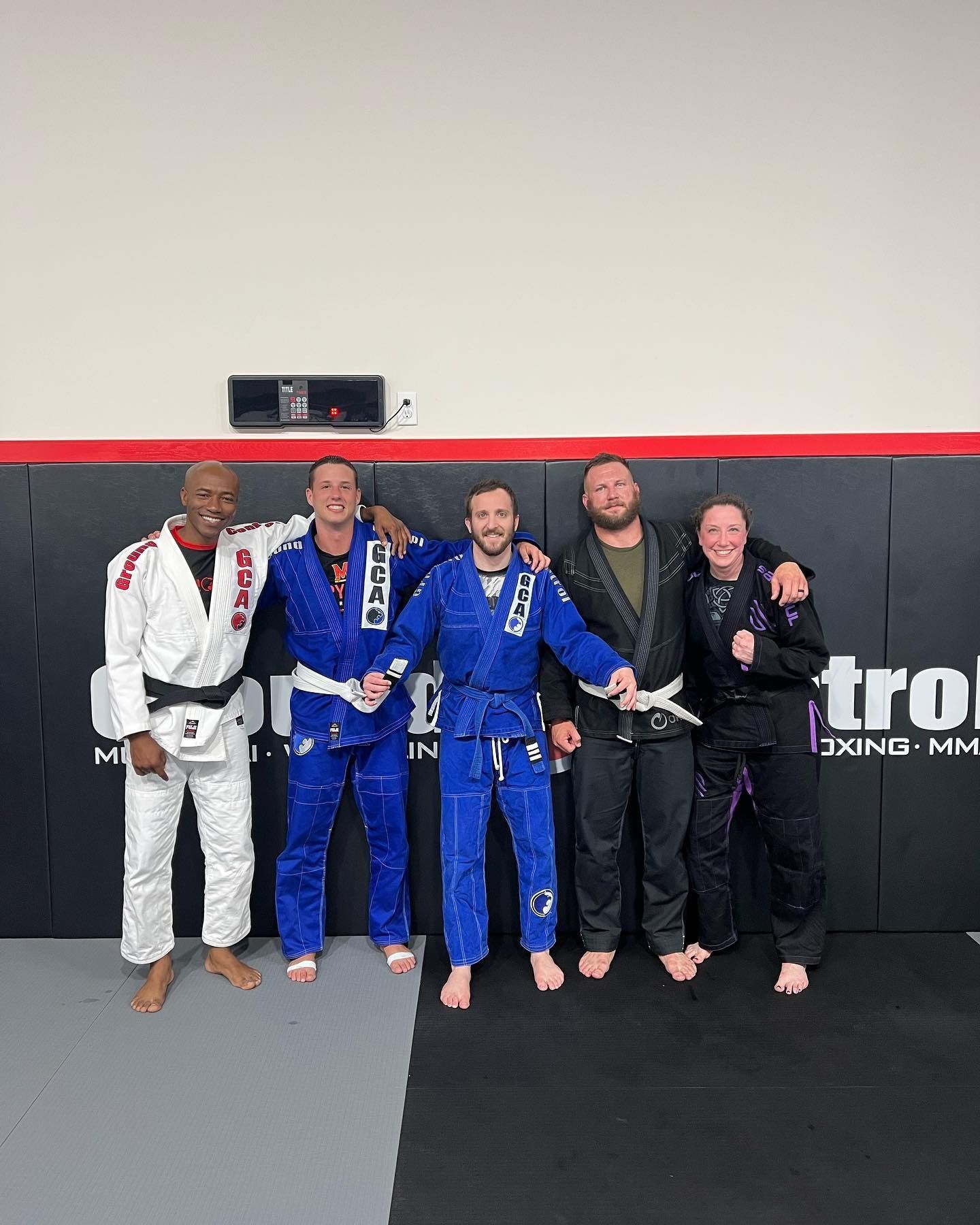 Five people in martial arts uniforms, posing in a gym with mats.