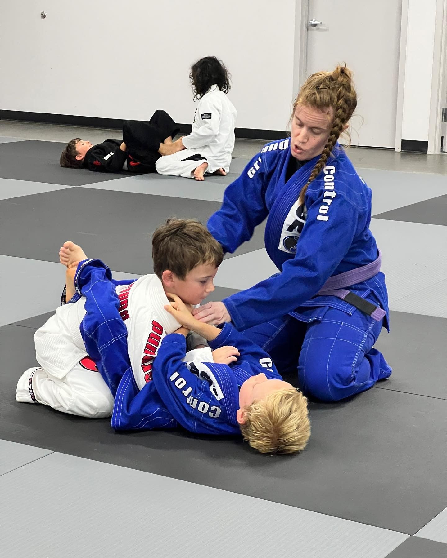 Woman in blue gi coaching two young boys in blue gis on a mat; other children watch.