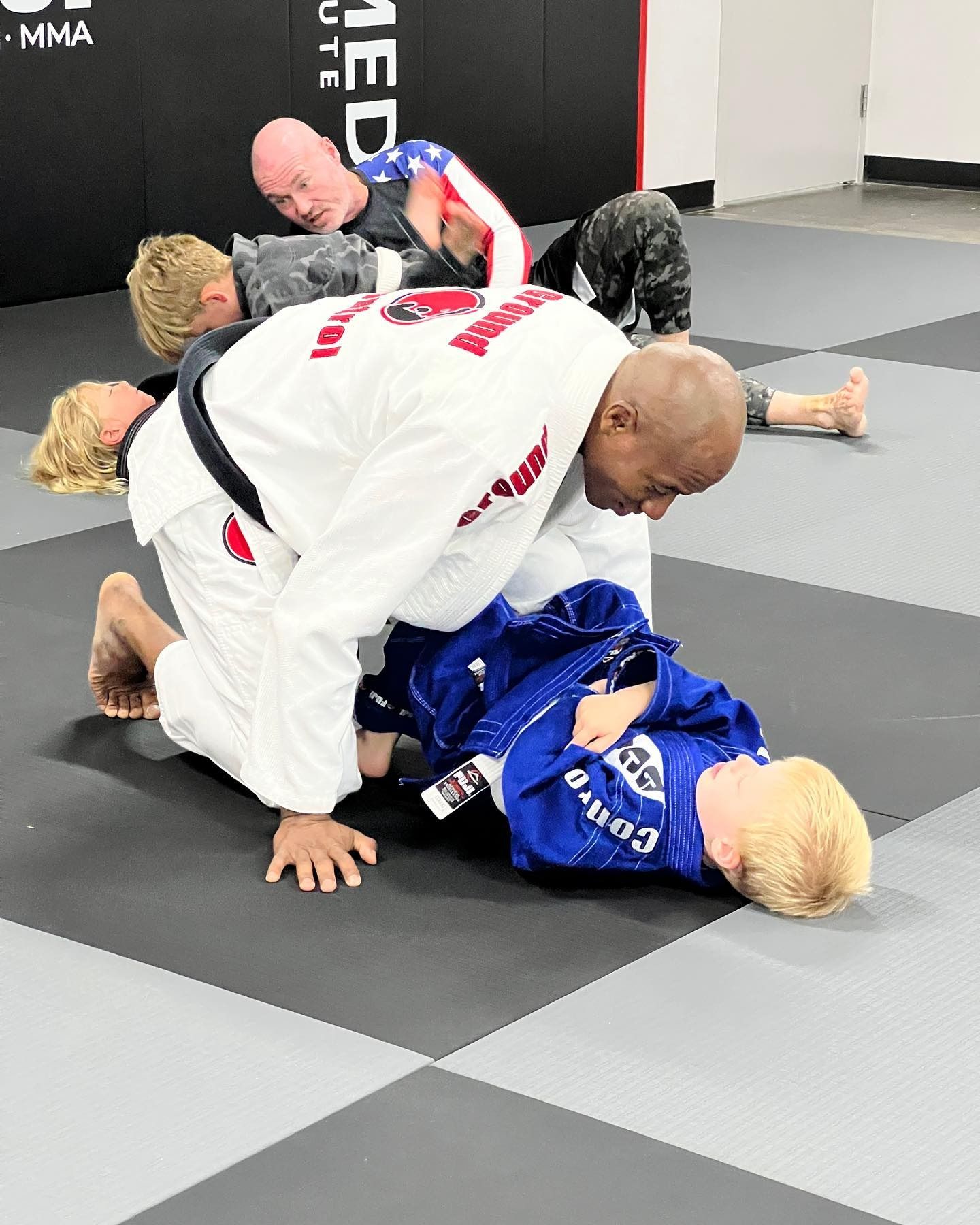 People practicing Brazilian Jiu-Jitsu. An instructor in a white gi kneels over a child in a blue gi on a mat.
