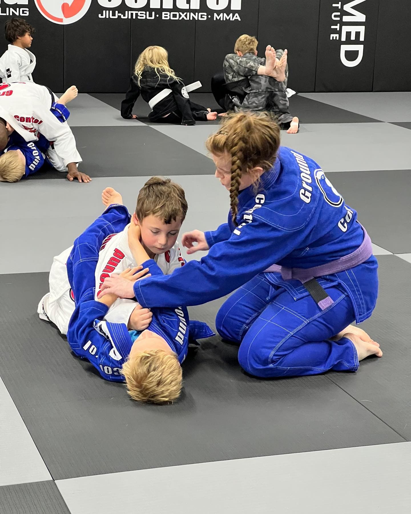 Two kids in blue gis practice Jiu-Jitsu with an instructor on a gray mat. Other students watch.