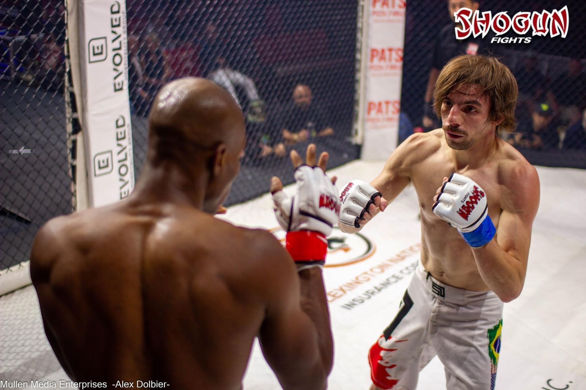 Two MMA fighters in a ring. One with a fro is punching, and the other is blocking. Fight is in a dark arena.