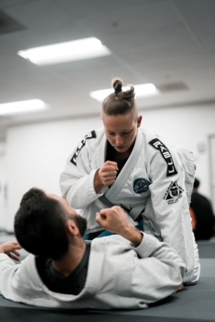 Woman in a white gi over a man on the floor, in a martial arts training session.