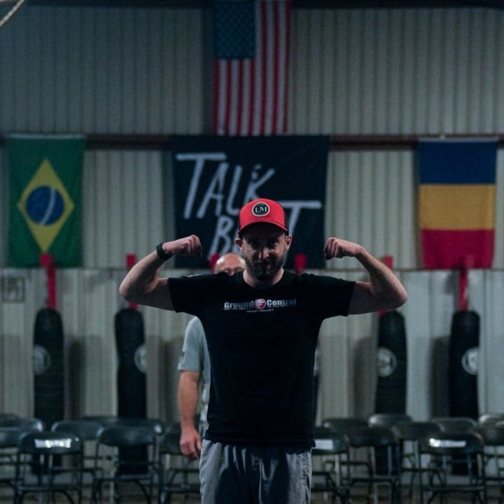 Man flexing in gym, wearing a red hat and black shirt, with flags of Brazil, USA, and Romania in the background.