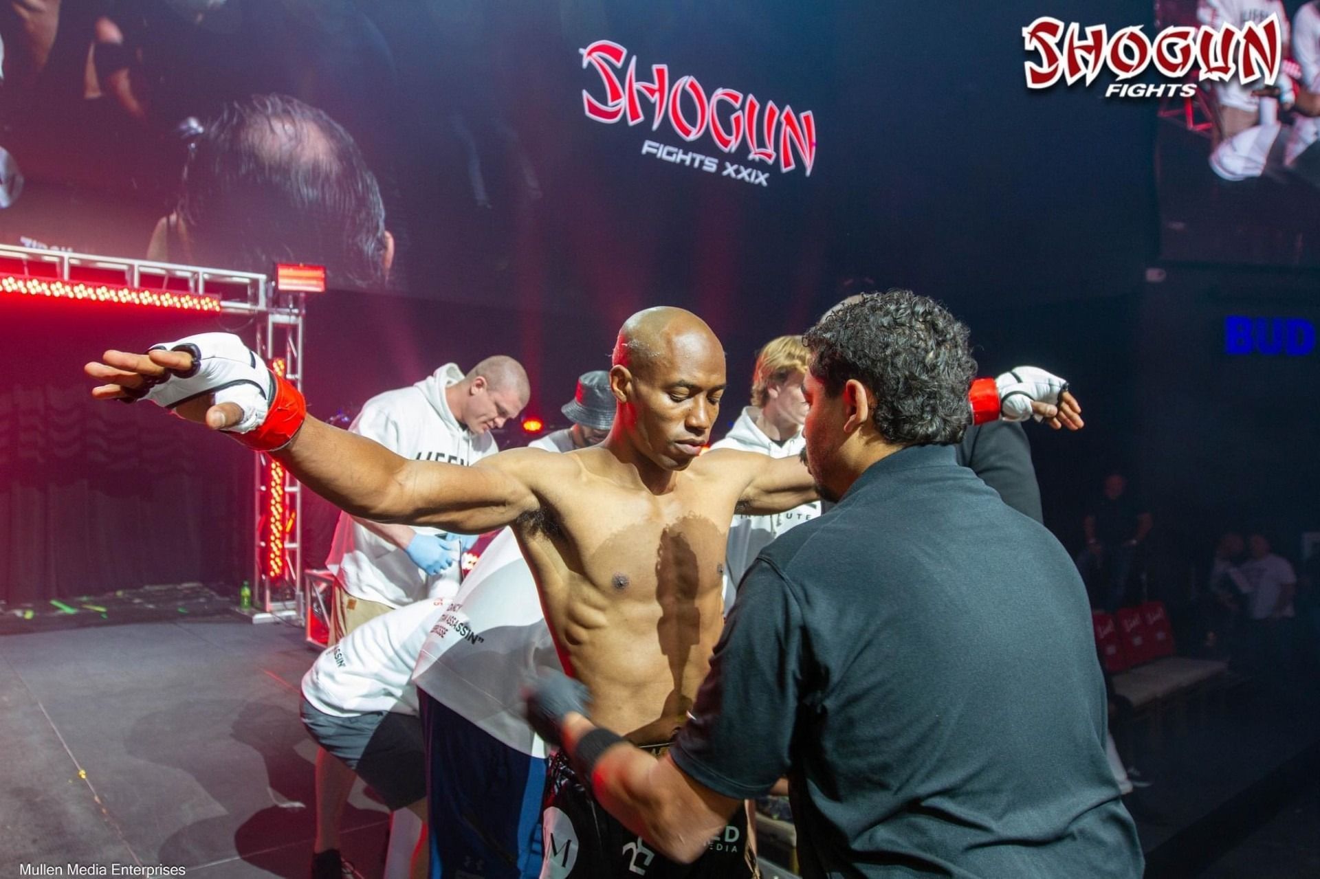 Male MMA fighter in a ring, arms outstretched, being checked by officials before a fight. 