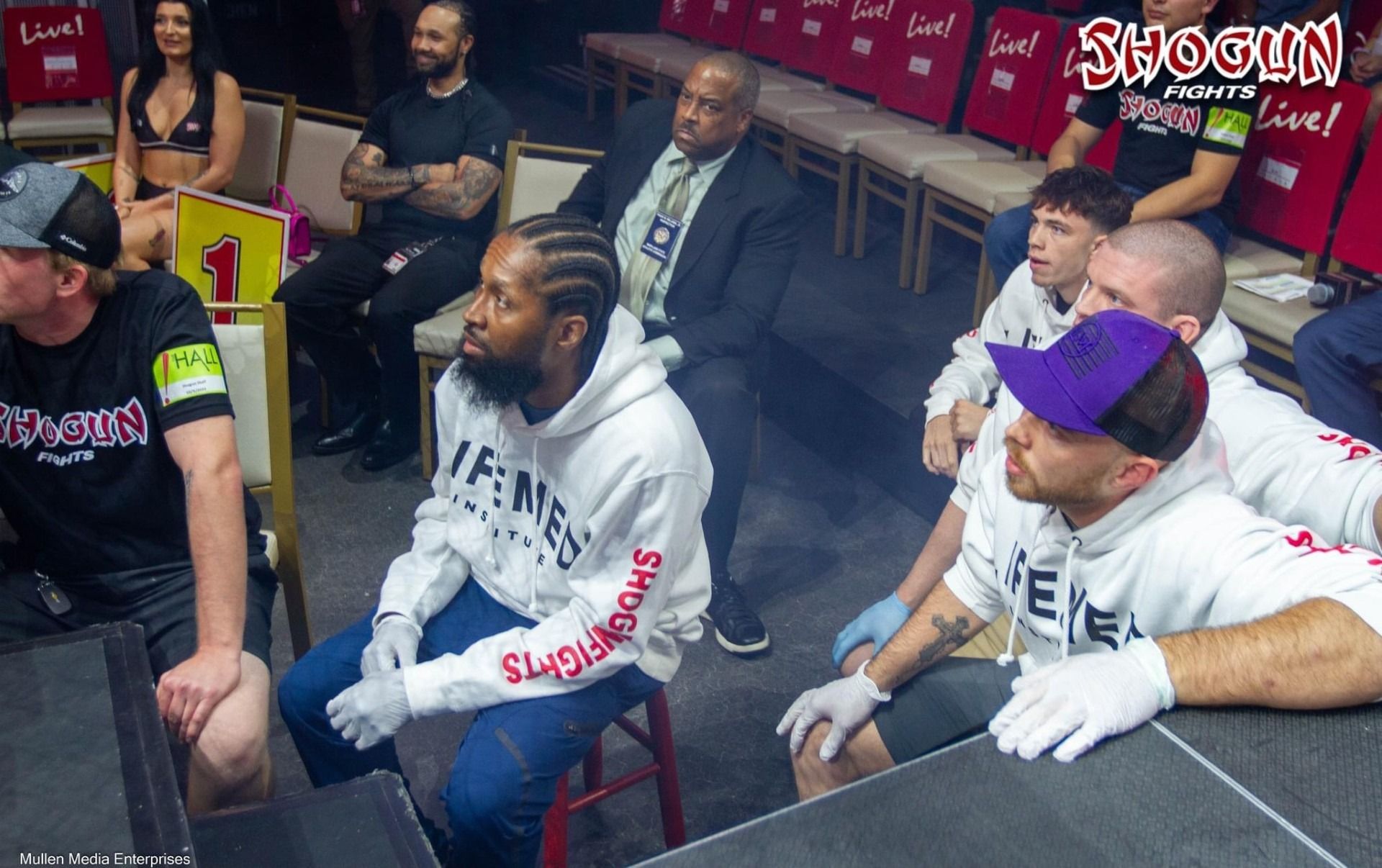 People in ringside seating at a fight: Black man in white hoodie, others watching with various expressions.