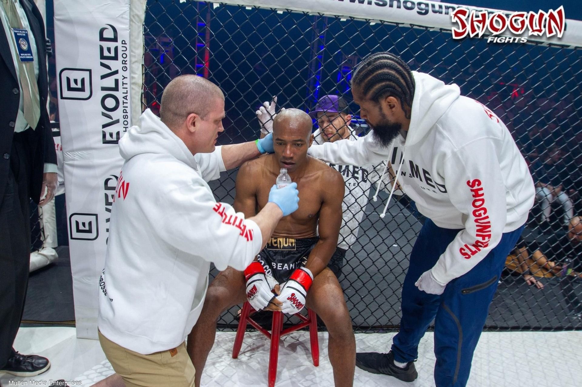 A fighter being attended to by two people in a cage. The fighter has a mouthpiece in his mouth, looking distraught.