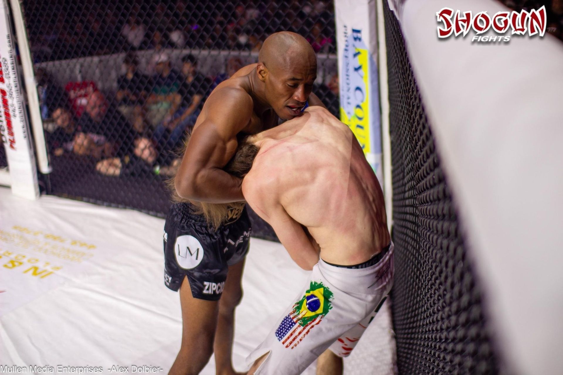 MMA fighters in a cage, one with dark skin, grappling the other with a white skin, leaning against the fence.