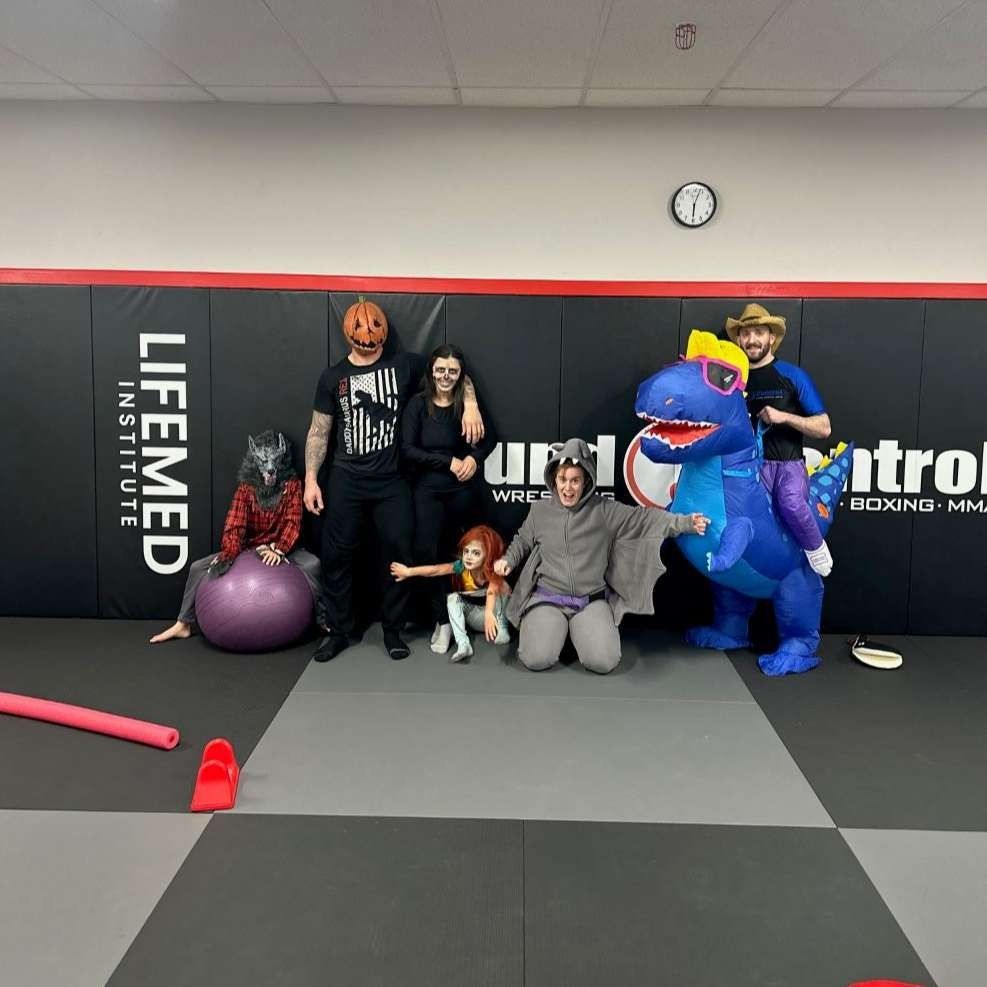 Group in Halloween costumes posing in a gym: pumpkin head, wolf, dinosaur, witch, cowboy, etc.