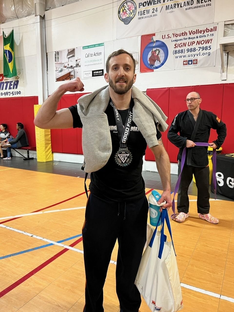 Man flexes with medal, towel, and bag at a jiu-jitsu tournament. Another man in a gi stands behind.