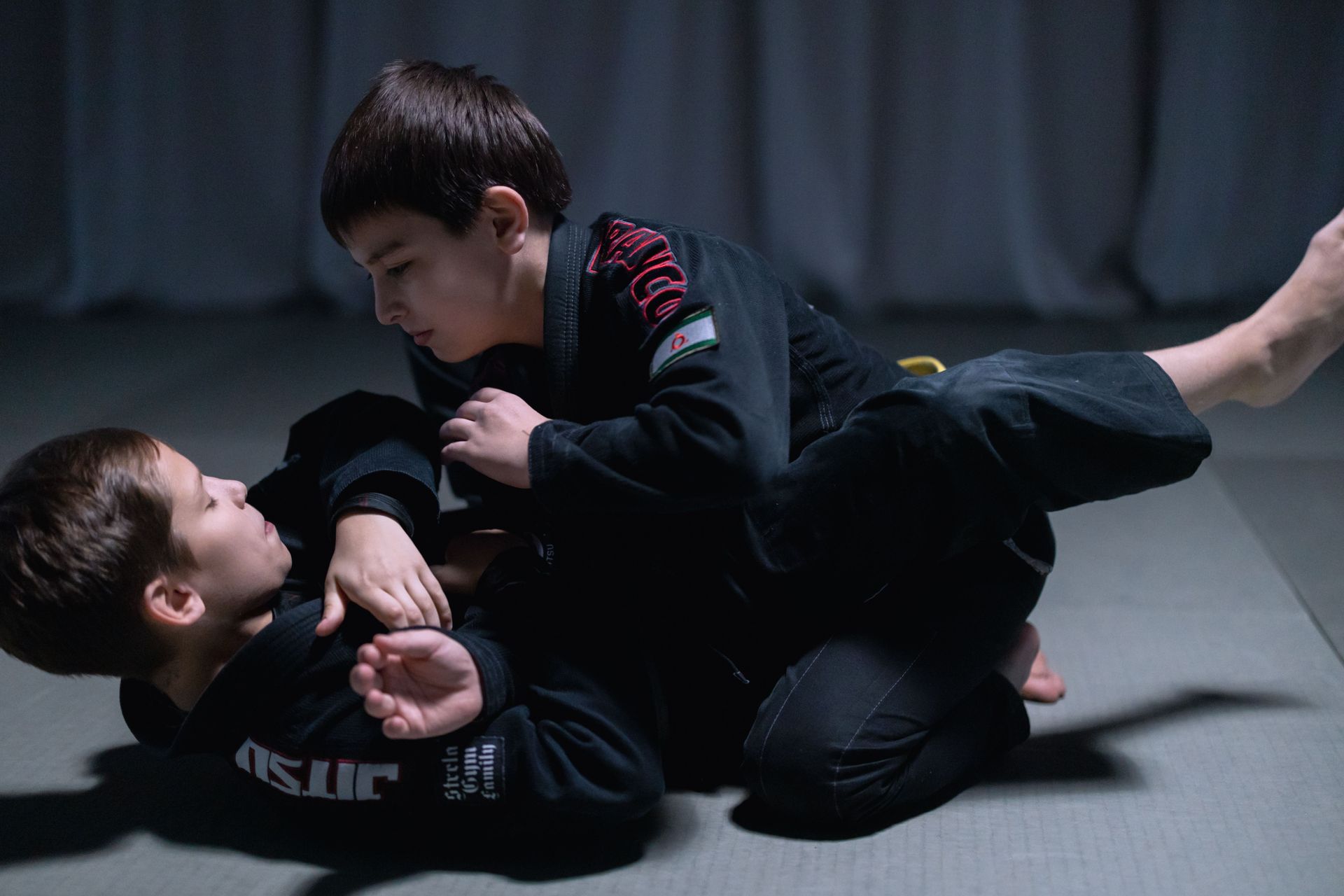 Two martial artists grappling on mats in a dojo, with two others blurred in the background.