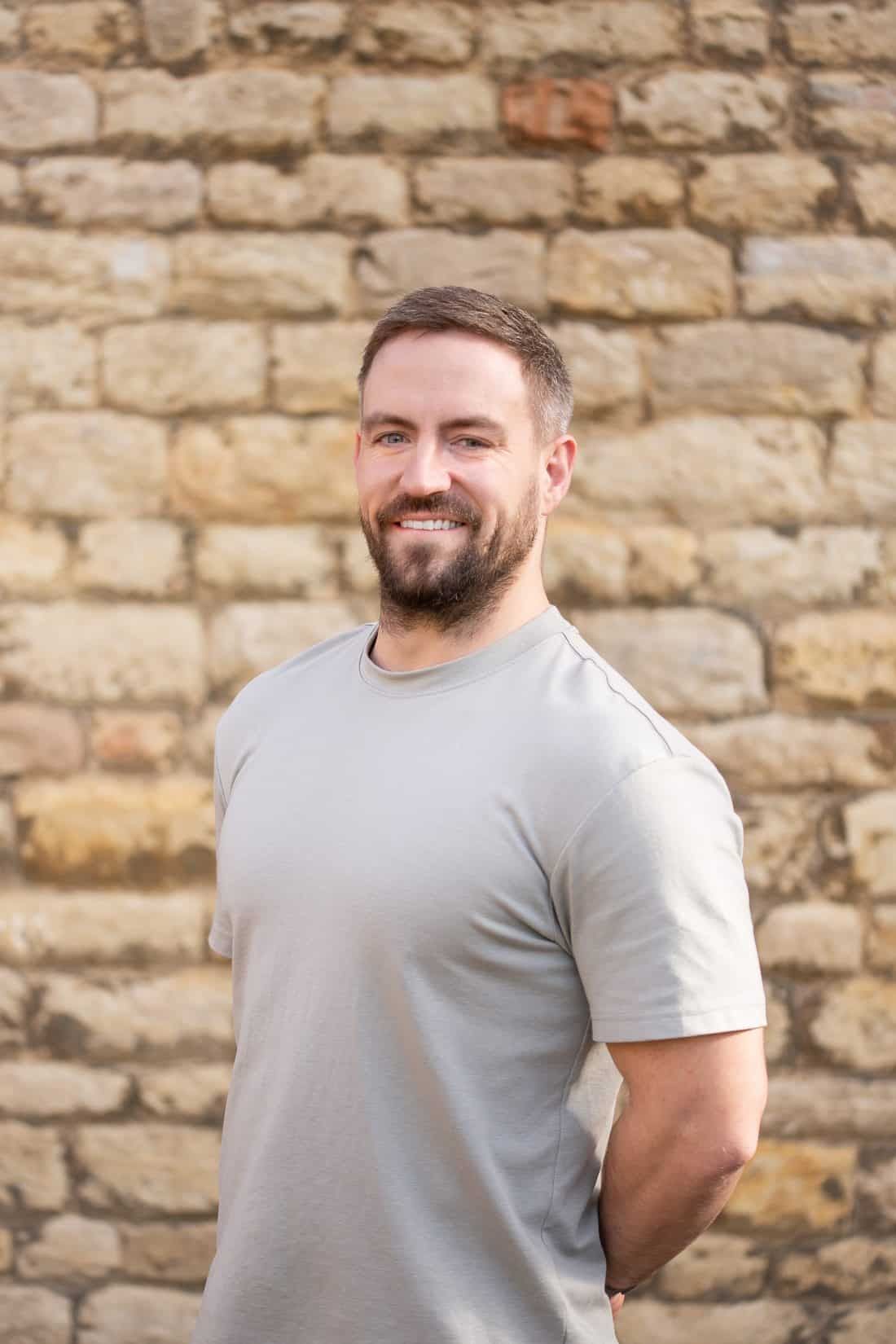 A person with a beard, wearing a grey t-shirt, smiling while standing in front of a textured stone wall.