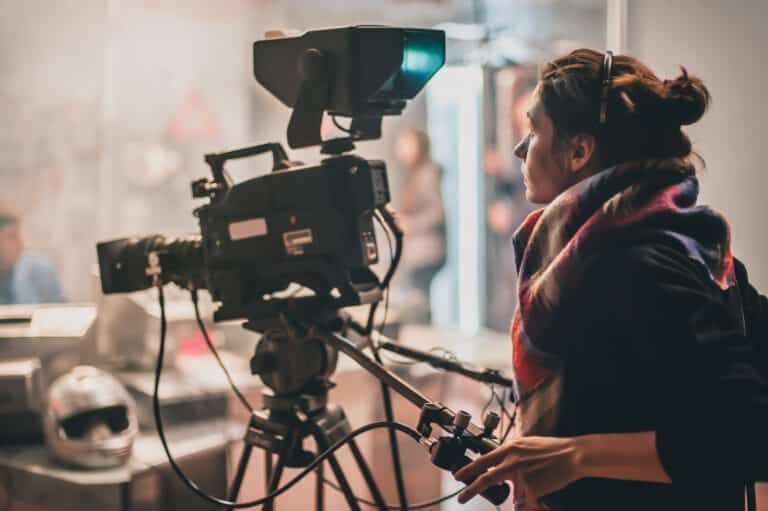 A person with their hair in a bun stands behind a professional video camera on a tripod in a dimly lit studio.
