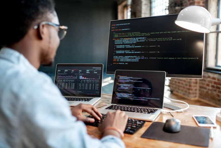 A person wearing glasses works at a desk, typing on a keyboard with a laptop and a large monitor displaying code.