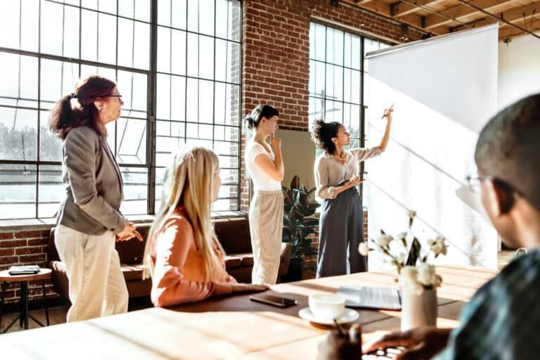 A small team collaborates in a sunlit office, with one person gesturing to a whiteboard during a presentation.