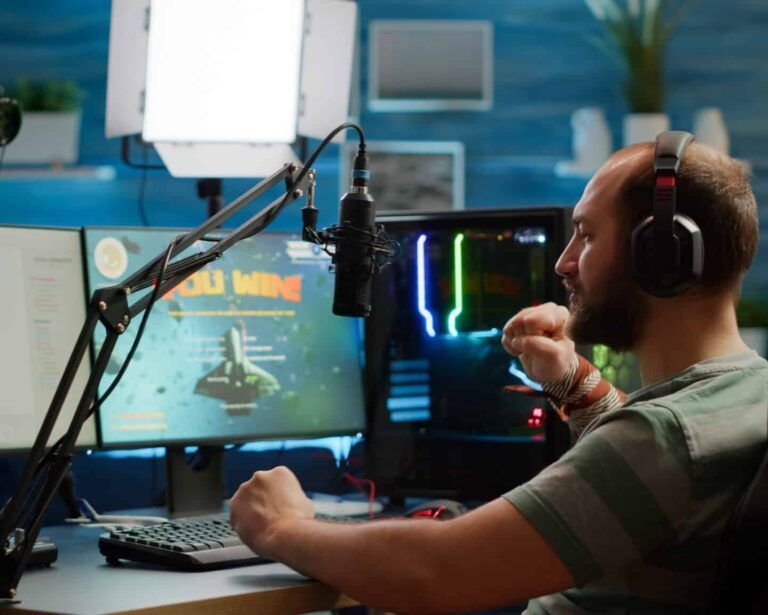 A streamer in headphones celebrates a gaming victory at a desk with a microphone, light panel, and computer equipment.