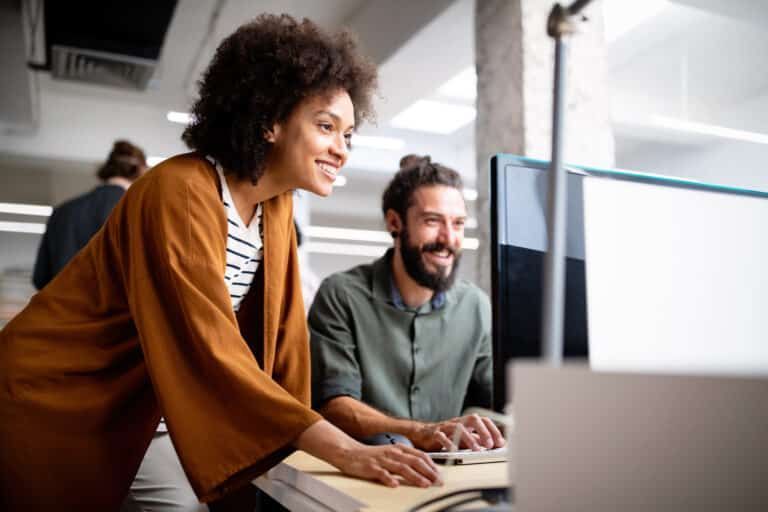 Two colleagues collaborating in an office, smiling while looking at a computer screen.