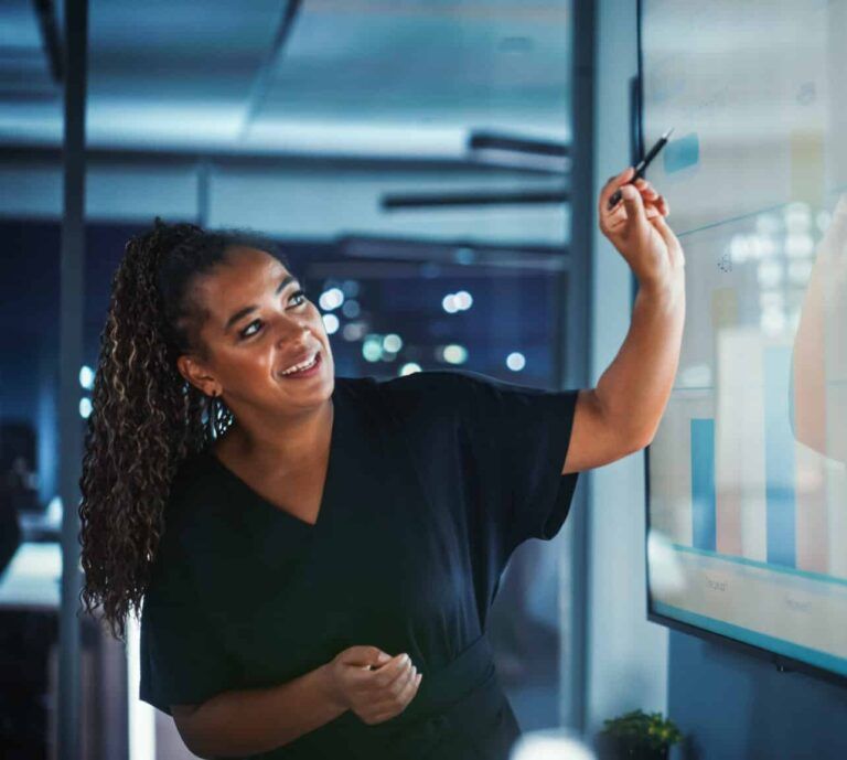 A professional points to a digital bar chart on a screen in a modern office at night.