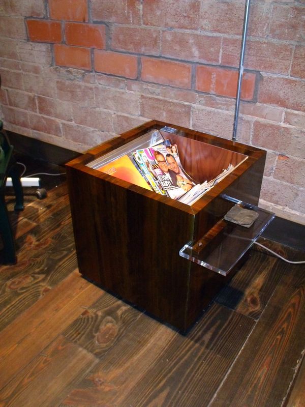 A wooden box filled with magazines sits on a wooden floor in front of a brick wall