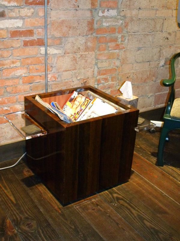 A wooden box filled with magazines sits on a wooden floor in front of a brick wall.