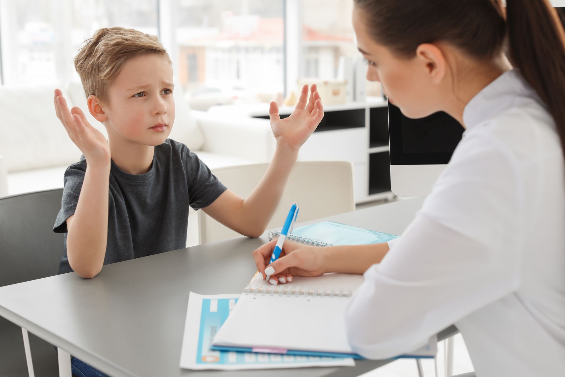 A young boy is sitting at a table talking to a woman.