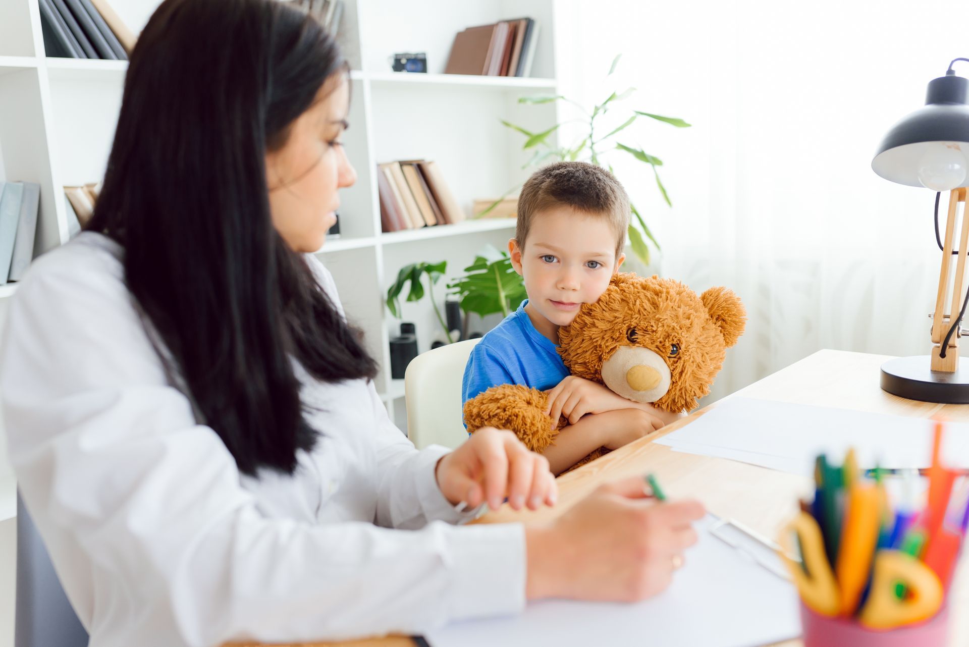 A woman is sitting at a table with a child holding a teddy bear.