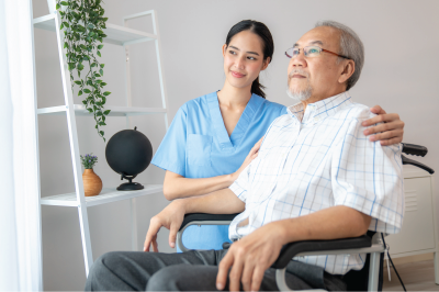 Caregiver comforts an elderly person in a wheelchair indoors, with a shelf in the background.