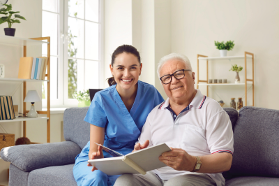 Woman in scrubs and elderly man looking at a photo album, smiling on a couch. Living room setting.