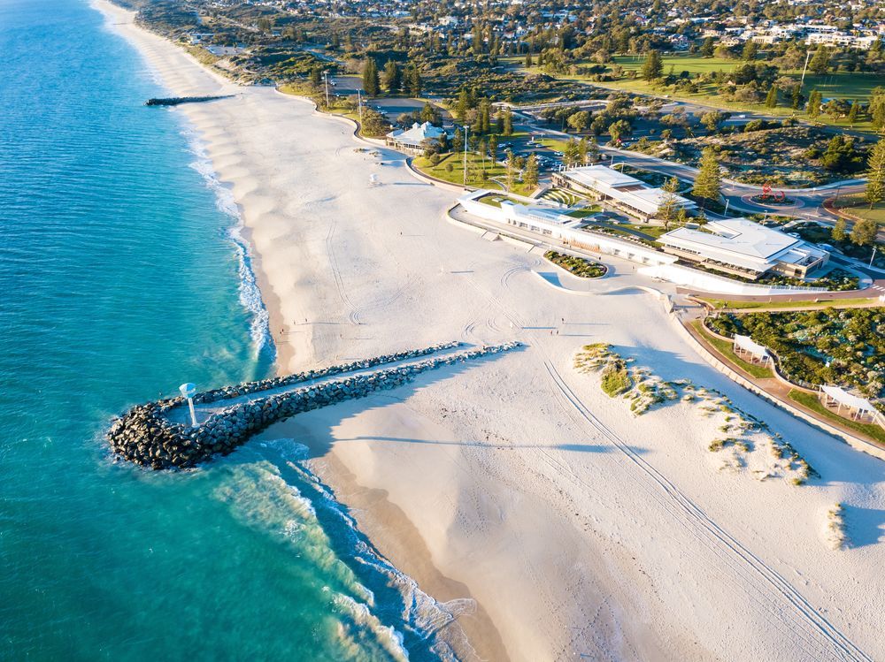 Aerial view of a white sand beach, turquoise water, and a stone jetty; buildings and greenery in the background.