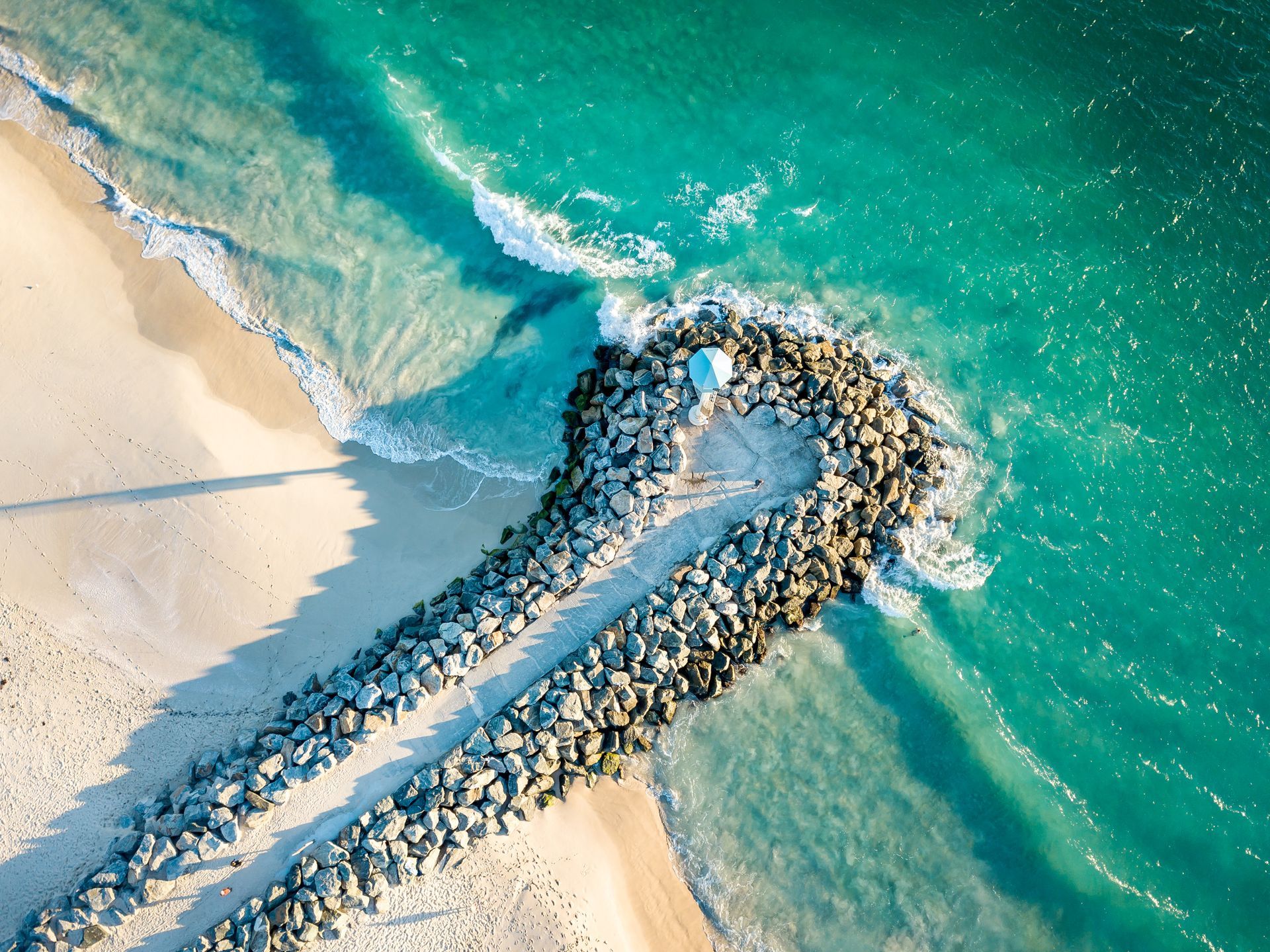 Aerial view of a white sand beach, turquoise water, and a stone jetty; buildings and greenery in the background.