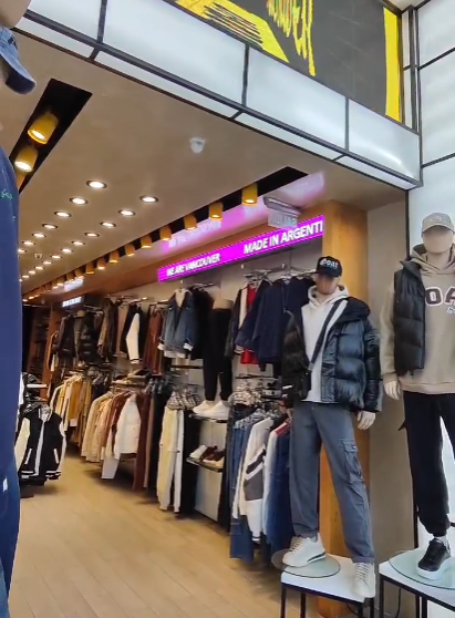 Interior de una tienda de ropa con maniquíes, percheros y letreros luminosos.