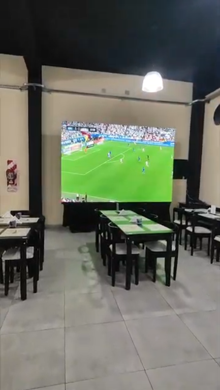 Interior de un comedor con una pantalla grande que muestra un partido de fútbol. Se ven mesas y sillas.
