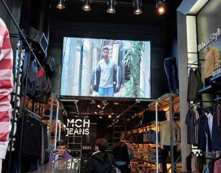 Interior de una tienda de ropa con una gran pantalla que muestra a una persona con chaqueta y vaqueros.