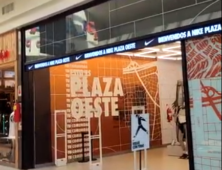 Exterior de una tienda Nike en Plaza Oeste, con letreros naranjas y blancos y una puerta de seguridad en la entrada.