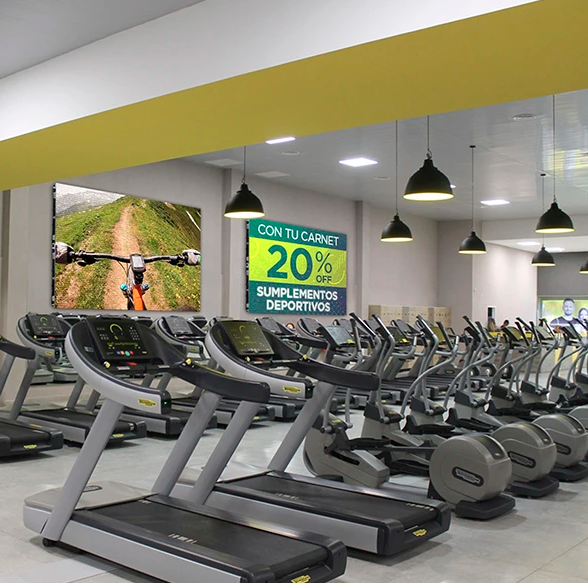 Interior del gimnasio con cintas de correr, máquinas elípticas y un cartel que ofrece un 20% de descuento en suplementos.