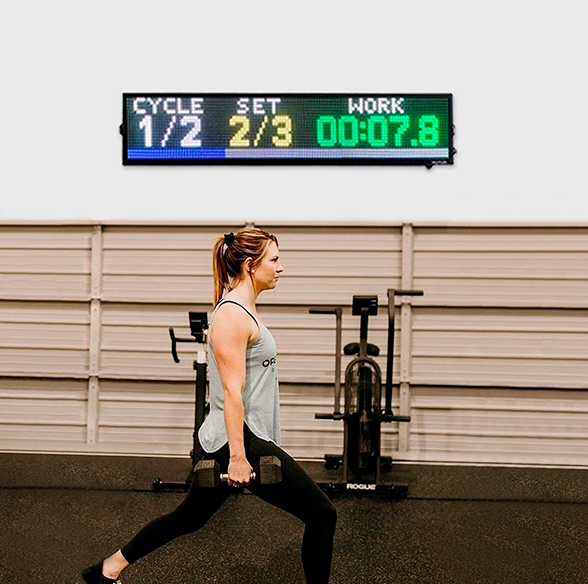 Una mujer realiza estocadas con mancuerna en el gimnasio bajo un cronómetro digital, contando series y tiempo.